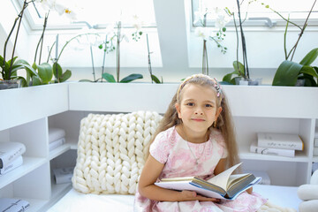 Banner with a cute Caucasian girl in a pink dress with a book sitting in the children's room. Reading and education. Children concept. Copy space on the left