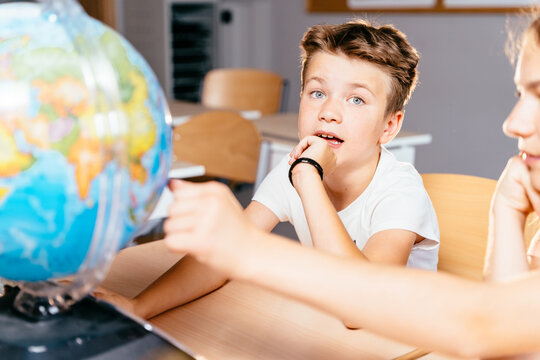 Cute Pupil Boy Inquiringly Looking At Camera Children In School Find Something At The Globe At Classroom At School. Make The World A Better Place.