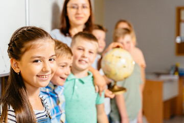 Group of school children with teacher standing in classroom. Portrait of primary schoolboys and schoolgirls standing in a line near chalkboard. Focus on cute girl.