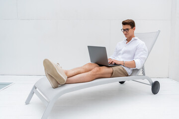 full length of smiling freelancer in summer clothes sitting in deck chair and typing on laptop.