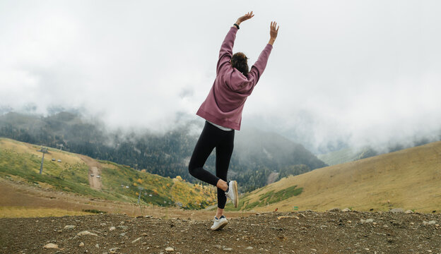 Hyped cheerful young woman jumping, stunned by view. On grassy mountains surrounded by low hanging clouds. She is wearing a pink hoodie.