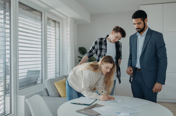 Young couple signing contract when buying their new home and receiving keys from real estate agent.