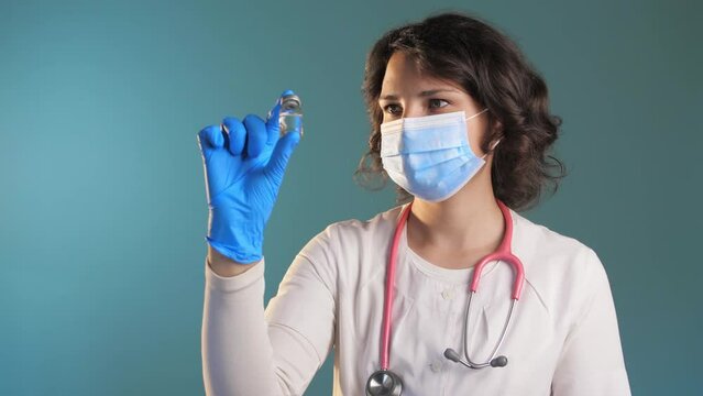 Woman doctor in a medical mask stands in a blue background and looks at a ampule of a vaccine.Transparent vial of a medical drug.Slow Motion