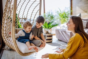 Young cheerful mother playing with her little children and having fun when swinging them on hanging chair in conservatory at home.