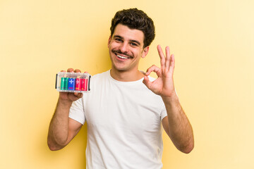 Young caucasian man holding batteries isolated on yellow background cheerful and confident showing ok gesture.