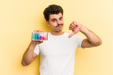 Young caucasian man holding batteries isolated on yellow background showing a dislike gesture, thumbs down. Disagreement concept.
