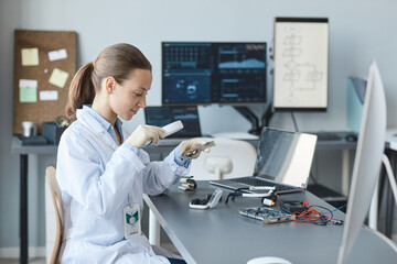 Side view portrait of young female scientist inspecting hardware with magnifying glass in...