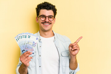 Young caucasian man holding banknotes isolated on yellow background smiling and pointing aside,...