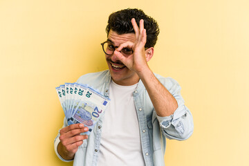 Young caucasian man holding banknotes isolated on yellow background excited keeping ok gesture on...