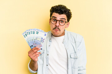 Young caucasian man holding banknotes isolated on yellow background shrugs shoulders and open eyes...