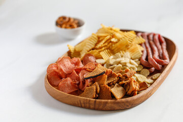 Wooden dish, plate with beer snacks, potatoes chips, meat chips, sausages kabanos. White background, copy space.