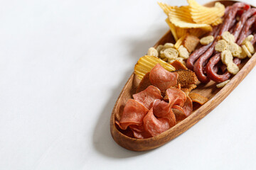 Wooden oak oval dish, plate with beer snacks, potatoes chips, meat chips, sausages kabanos, bread crackers. White background, copy space.
