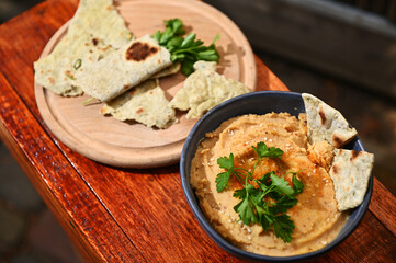Overhead view of a bowl of chickpea hummus and a wooden board with parsley wrapped in pit bread, on a wooden table