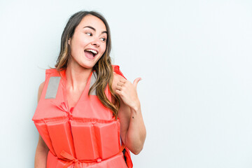 Young caucasian woman wearing life jacket isolated on blue background points with thumb finger...