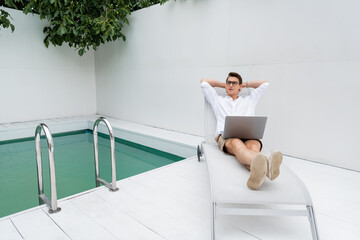 full length of man with laptop relaxing in deck chair near pool.