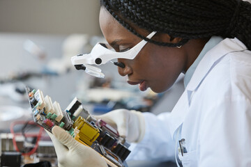Side view portrait of black female scientist wearing magnifying glasses and inspecting electronic...