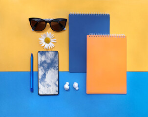 Attributes of  blogger in summer -  mobile phone with  reflection of the sky,  pen, headphones, notebooks, sunglasses, daisies, top view, colorful bright background