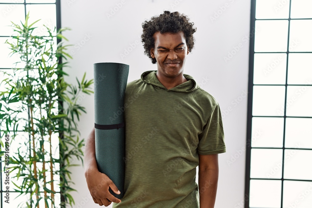 Poster african man with curly hair holding yoga mat at studio winking looking at the camera with sexy expre