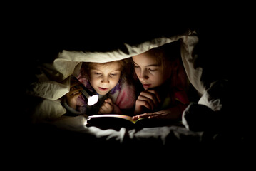 two sisters are reading a book under the covers at night with a flashlight - obrazy, fototapety, plakaty