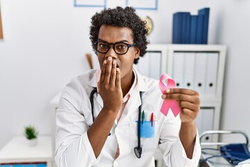 African doctor man holding pink cancer ribbon covering mouth with hand, shocked and afraid for...