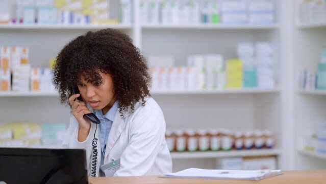Friendly pharmacist talking on a phone call and checking something on her computer in a pharmacy. Woman using pc to access drug database for inventory check or access customers prescription online