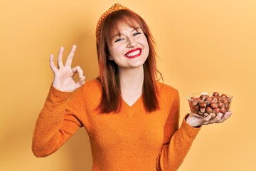 Redhead young woman holding hazelnuts doing ok sign with fingers, smiling friendly gesturing excellent symbol