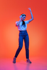 Fototapeta premium Full-length portrait of young pretty girl, student in casual style clothes isolated on orange color background in neon light. Concept of beauty, fashion, emotions