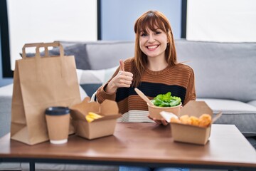 Obraz na płótnie Canvas Young beautiful woman eating delivery food at the living room smiling happy and positive, thumb up doing excellent and approval sign
