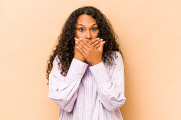 Young hispanic woman isolated on beige background covering mouth with hands looking worried.