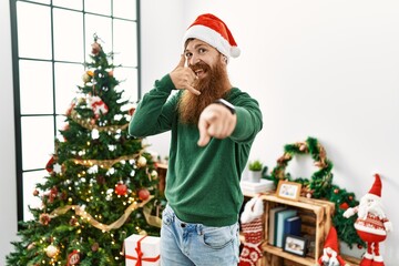 Redhead man with long beard wearing christmas hat by christmas tree smiling doing talking on the telephone gesture and pointing to you. call me.