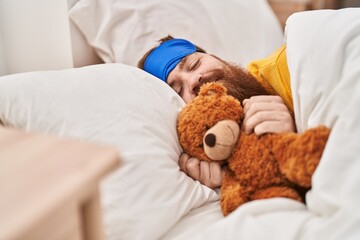 Young redhead man lying on bed hugging teddy bear at bedroom