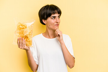 Young caucasian man holding crisps isolated on yellow background looking sideways with doubtful and skeptical expression.