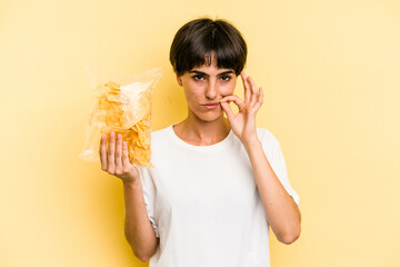 Young caucasian man holding crisps isolated on yellow background with fingers on lips keeping a secret.