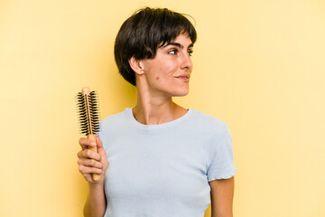 Young caucasian woman holding a brush hair isolated on yellow background looks aside smiling, cheerful and pleasant.