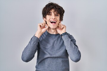 Young man standing over isolated background smiling pulling ears with fingers, funny gesture....