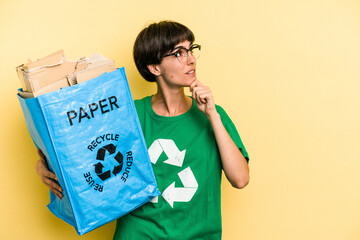 Young woman holding a recycling bag full of paper to recycle isolated on yellow background looking sideways with doubtful and skeptical expression.