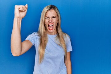 Fototapeta premium Beautiful blonde woman wearing casual t shirt over blue background angry and mad raising fist frustrated and furious while shouting with anger. rage and aggressive concept.