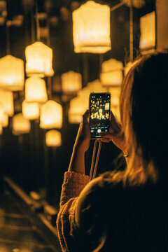 Woman Photographing Yellow Floating Lamps With Smartphone In Dark Room