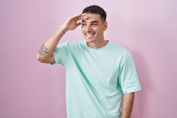 Handsome hispanic man standing over pink background very happy and smiling looking far away with hand over head. searching concept.