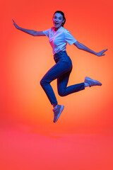 Fototapeta na wymiar Full-length portrait of young pretty girl, student in casual style clothes isolated on orange color background in neon light. Concept of beauty, fashion, emotions