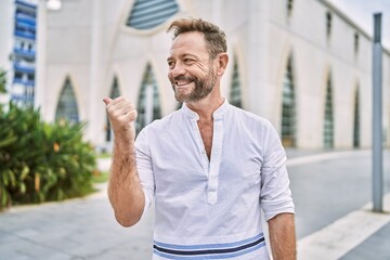 Fototapeta premium Middle age man outdoor at the city smiling with happy face looking and pointing to the side with thumb up.