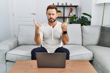 Middle age man using laptop at home pointing up looking sad and upset, indicating direction with...