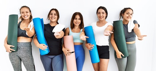 Group of women holding yoga mat standing over isolated background smiling cheerful offering palm hand giving assistance and acceptance.