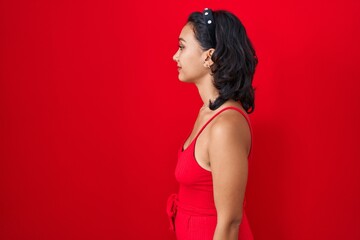 Young hispanic woman standing over red background looking to side, relax profile pose with natural face with confident smile.