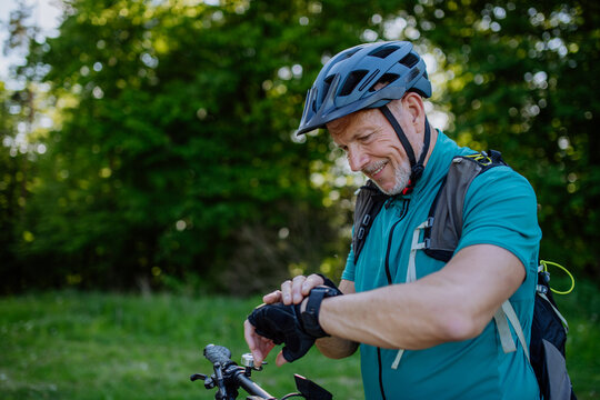 Active senior man riding bicycle at summer park, looking at smartwatch and checking his performance, healthy lifestyle concept. - Powered by Adobe