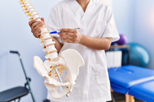 Young latin woman wearing physiotherapist uniform pointing to anatomical model of vertebral column at physiotherapy clinic
