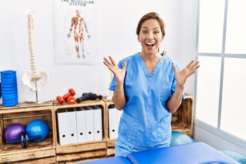 Middle age hispanic physiotherapist woman working at pain recovery clinic celebrating crazy and amazed for success with arms raised and open eyes screaming excited. winner concept