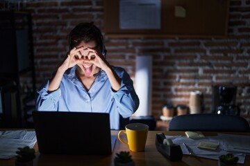 Beautiful brunette woman working at the office at night doing ok gesture like binoculars sticking tongue out, eyes looking through fingers. crazy expression.