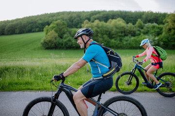 Active senior couple riding electric bicycles on trail at summer park, healthy lifestyle concept. Side view.