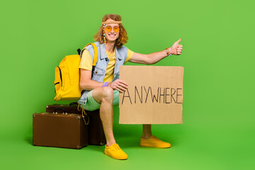 Full size photo of positive hippy person sitting suitcase thumb up hitchhike isolated on green color background